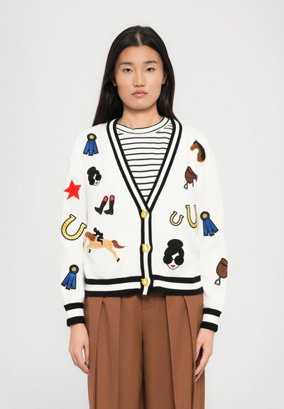 Jeune femme portant un cardigan blanc avec des bordures noires et des patchs sur le thème équestre par-dessus une chemise à rayures noir et blanc et un pantalon marron.