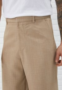 Man som bär beigefärgade rutiga byxor och vit skjorta, med en tatuerad högra underarm synlig mot en vit tegelvägg som bakgrund.