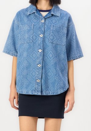 Henkilö, jolla on lyhythihainen sininen kuvioitu farkkupaita, jossa on edessä napit, mustan hameen päällä, yksiväristä valkoista taustaa vasten.