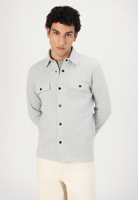 Veste grise claire à boutons, en tissu texturé, dotée de deux poches poitrine avec des boutons noirs et d'un col classique.