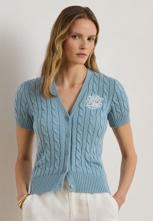 Kobieta ubrana w jasnoniebieski krótkorękawowy sweter z warkoczowym splotem, zapinany na guziki, ozdobiony białymi haftowanymi inicjałami, w połączeniu z białymi spodniami i złotymi bransoletkami.