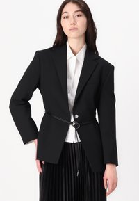 Black blazer with a fitted silhouette, single button, and a contrasting black belt at the waist. Worn over a white button-up shirt.