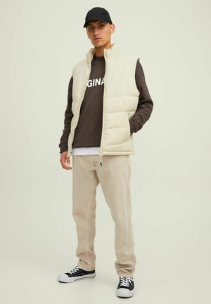 Beige puffer vest over a brown sweatshirt with "ORIGINAL" printed. Light-colored pants and black sneakers with white soles. Black cap.