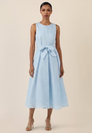 Woman standing wearing a sleeveless light blue textured midi dress with a front bow tie at the waist and beige high-heeled sandals.