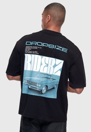 Bărbat purtând un tricou negru cu o mașină decapotabilă vintage albastră și un text mare "RIDERZ" printat pe spate.