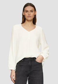 Blouse blanche texturée à manches longues avec un décolleté en V et une fermeture à boutons. Assortie à un jean foncé, le modèle pose sur un fond neutre.
