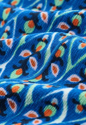 Modrá látka s texturovaným povrchem, vyznačující se květinovým vzorem v odstínech zelené, oranžové a černé. Zakřivené vlny zvýrazňují hloubku a pohyb.