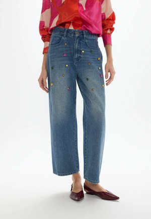 Mujer con vaqueros azules de talle alto con adornos de gemas de colores, una blusa de manga larga rosa y roja con estampado, y zapatos planos puntiagudos de color rojo oscuro.