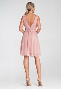 Hellrosa Kleid mit tiefem V-Ausschnitt auf der Rückseite und gebundenem Taillendetail. Fließender, transparenter Stoff reicht bis zur Mitte des Oberschenkels und wird mit flachen Schuhen mit Knöchelriemen kombiniert.