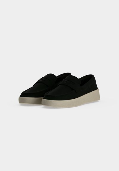 Paire de chaussures noires à enfiler avec semelles blanches épaisses présentée sur un fond uni gris clair.
