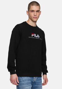 Musta huppari pitkillä hihoilla, pyöreällä kauluksella ja ribbireunoilla. Etupuolella on punavalkoinen brodeerattu "FILA"-logo.