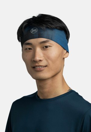 Joven con cabello corto y negro que lleva una diadema azul con estampado y una camisa azul marino, sonriendo ligeramente sobre un fondo blanco.