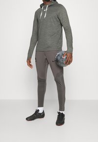 Hoodie gris à manches longues avec des cordons blancs, associé à un pantalon de sport gris ajusté présentant des accents texturés et des chaussures à crampons noires. Tient une balle grise.