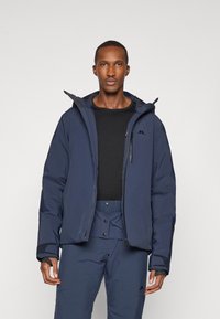 Casaco isolante azul-marinho com capuz, fecho com zíper e punhos ajustáveis. Feito com material liso e resistente à água, combinado com calças escuras.