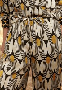 Pantalon en tissu avec un motif géométrique composé de lignes noires, d'un fond crème et de grands pois couleur moutarde. Détail de taille nouée visible.