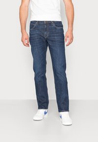 Jeans de mezclilla azul oscuro con corte recto, cinco bolsillos, costuras en contraste y una textura suave. Combinados con zapatillas blancas.