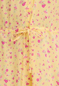 Tissu jaune avec petit motif floral rose et rouge, comprenant un bouton et une ceinture nouée à la taille en même matière.