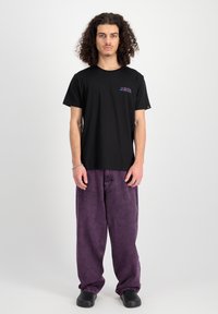 Schwarzes Baumwoll-T-Shirt mit kurzen Ärmeln und kleinem, mehrfarbigem "ALPHA INDUSTRIES"-Logo. Kombiniert mit lockeren, lila Jeanshosen und schwarzen Schuhen.