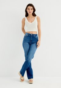 Top de punto blanco tipo cropped, jeans acampanados azules y sandalias de plataforma beige. El top presenta un tejido suelto y abierto; los jeans tienen un ajuste de altura media.