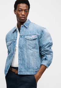 Muž se složenýma rukama, který má na sobě světle modrou džínovou bundu s vzorem přes bílou košili a tmavé kalhoty s hnědým opaskem.