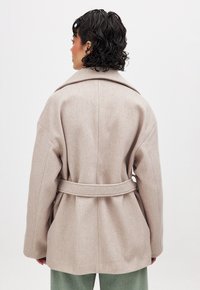 Abrigo de mezcla de lana beige con cintura ceñida, solapas anchas y hombros caídos. La textura parece suave, con un corte holgado y un diseño limpio.
