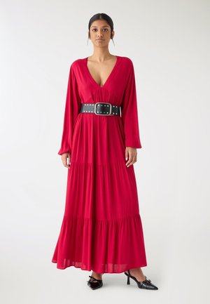 Donna che indossa un lungo vestito rosso con scollo a V profondo, cintura larga nera con borchie e décolleté nere con punta affilata, in piedi contro uno sfondo bianco.