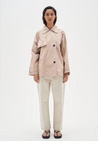 Vesimärkä beige trench coat, jossa on suuri kaulus ja kaksi mustaa nappia, yhdistettynä vaaleisiin kapeisiin housuihin ja mustiin sandaaleihin.