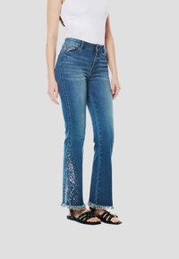 Széles szárú, kék farmer rojtos szegéllyel, finom fehér hímzéssel az oldalsó paneleken. Fekete pántos szandállal viselve.