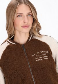 Brun og creme jakke lavet af tekstureret fleece, med lynlås foran og stribet krave. Har broderet tekst i hvid på den brune panel.