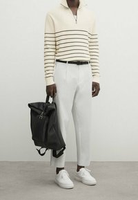 Pull-over blanc à rayures avec col à glissière, pantalon blanc, ceinture noire et sac à dos noir. Des baskets blanches complètent la tenue.