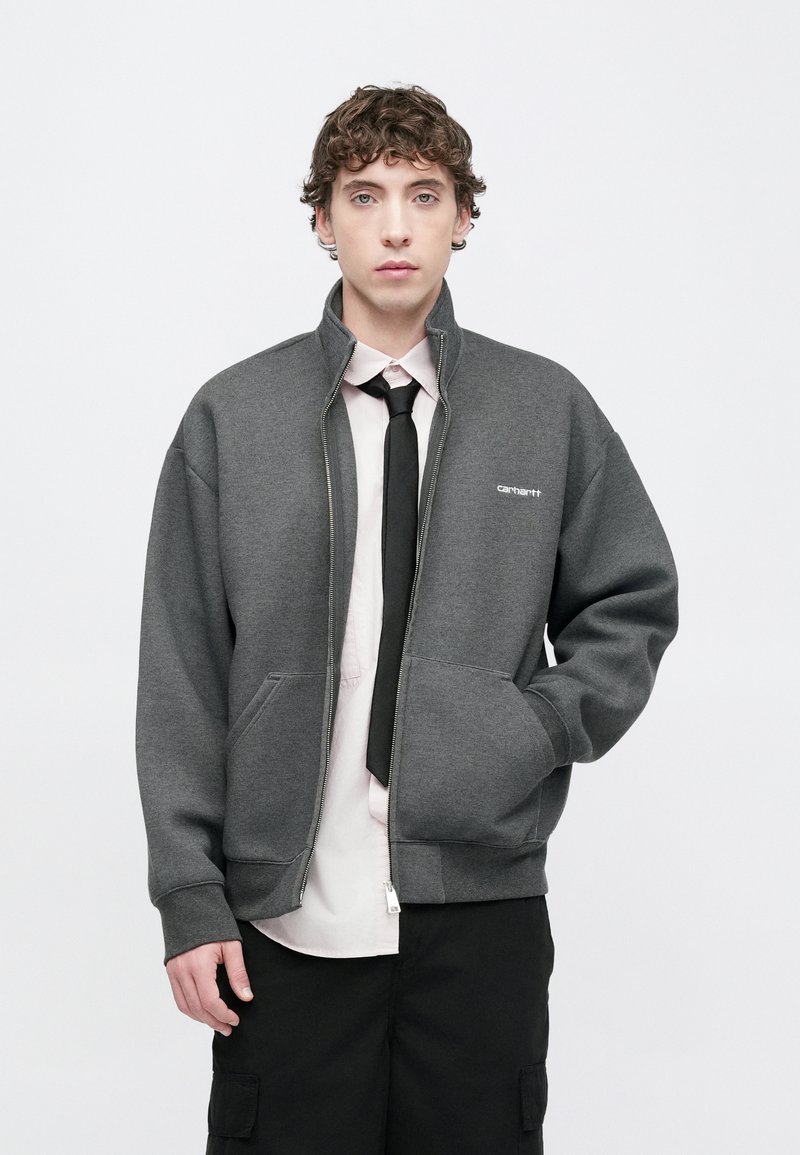 Sweat-shirt gris à fermeture éclair avec col montant, deux poches avant et détail logo blanc. Porté par-dessus une chemise rose clair et une cravate noire.