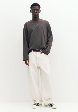 Jeune homme portant une chemise grise à manches longues, un pantalon blanc à jambes larges, une ceinture noire, un collier chaîne argenté et des baskets noires et blanches, debout.