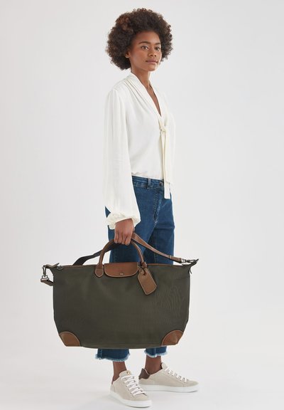 Olivgrüne Reisetasche aus Canvas mit braunen Lederelementen. Verfügt über zwei Griffe und einen verstellbaren Schultergurt. Glatte Textur.