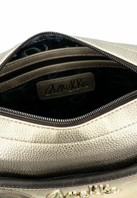 Goldfarbener Handtasche, die sich öffnet und ein dunkles Innenfutter sowie ein Etikett mit der Aufschrift "Anekke" im Reißverschlussfach zeigt.