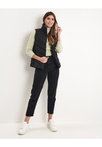 Gilet trapuntato nero sopra un maglione verde chiaro, abbinato a jeans neri a vita alta e sneakers bianche. La modella posa davanti a uno sfondo neutro.