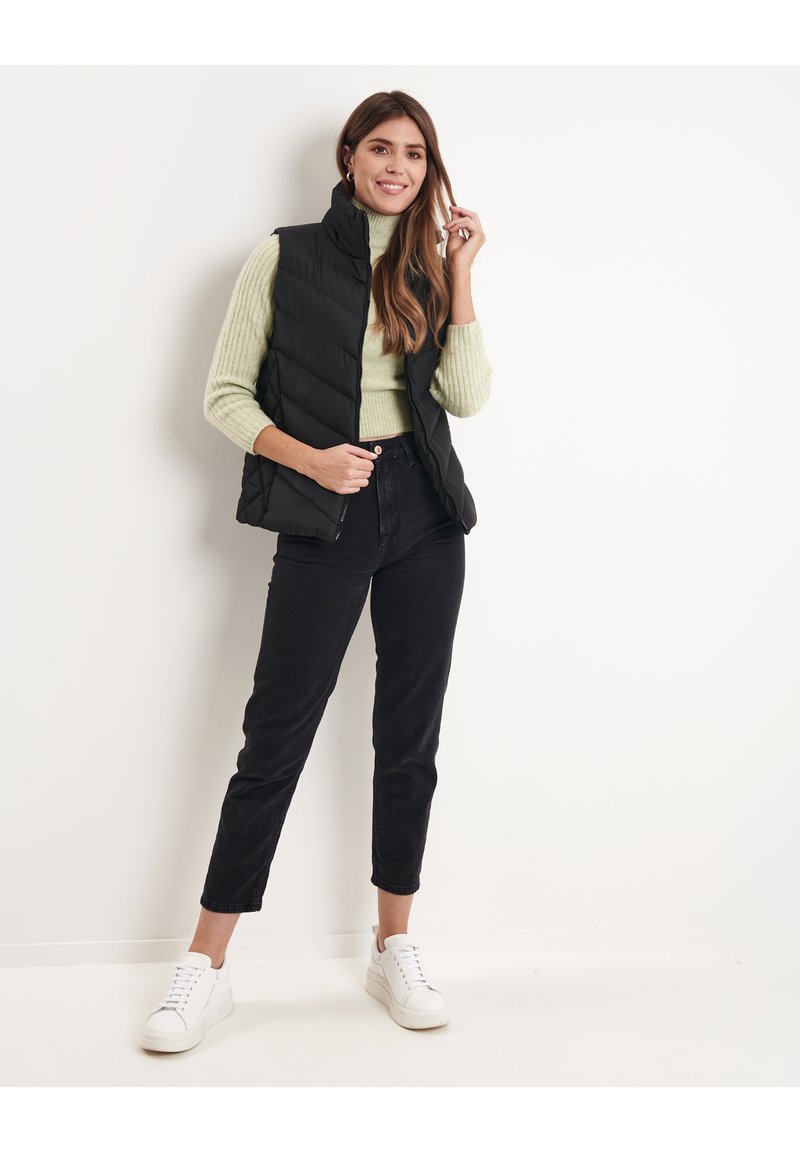 Gilet trapuntato nero sopra un maglione verde chiaro, abbinato a jeans neri a vita alta e sneakers bianche. La modella posa davanti a uno sfondo neutro.