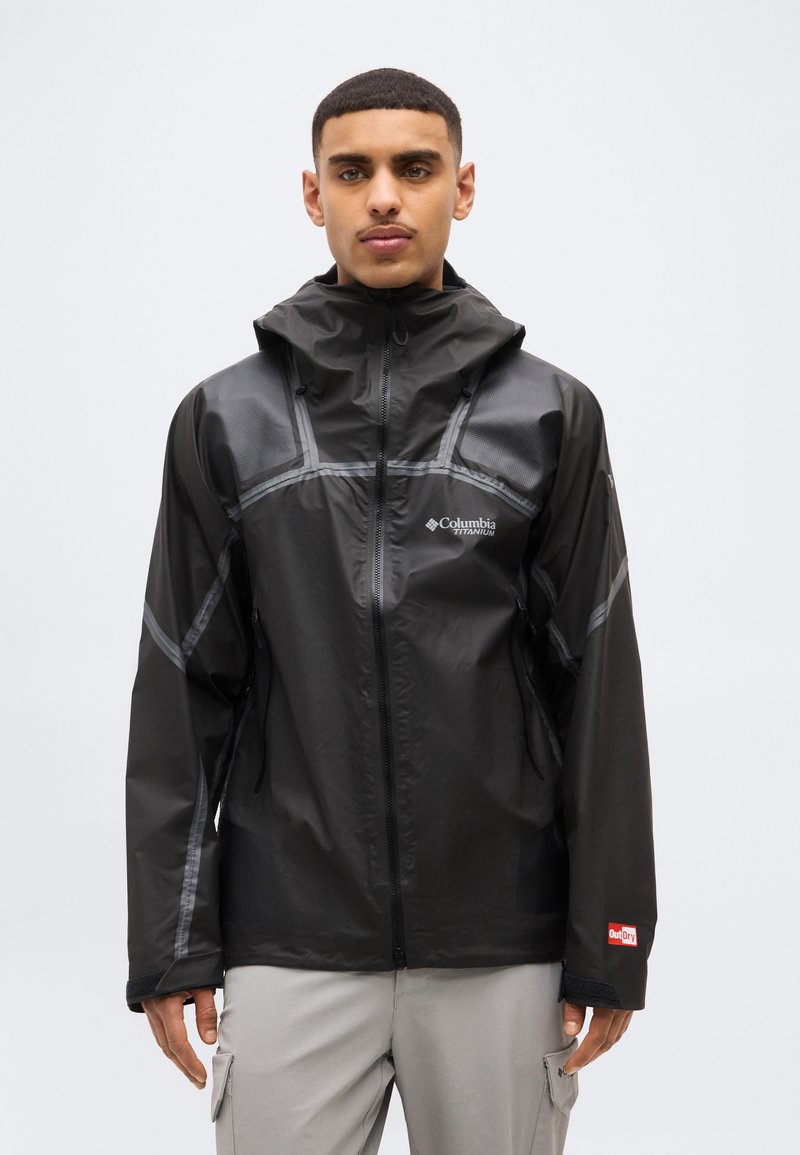 Νέος άνδρας που φοράει μαύρο αδιάβροχο μπουφάν Columbia Titanium με κουκούλα, στέκεται μπροστά σε απλό ανοιχτό φόντο.