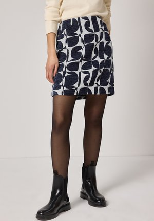 Femme portant une mini-jupe noire à motifs, des collants noirs transparents et des bottines en cuir noir, debout devant un fond clair.