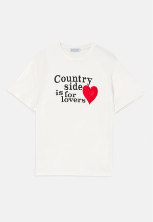 T-shirt de algodão branca com mangas curtas. Apresenta texto preto a dizer "O campo é para os amantes" e um gráfico de coração vermelho. Design simples.