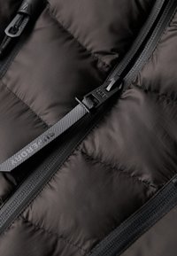 Sort puffervest med quiltet design, tekstureret stof og en plastiklynlås med en mærket trækstrop. Nærbillede fremhæver materialedetaljer.