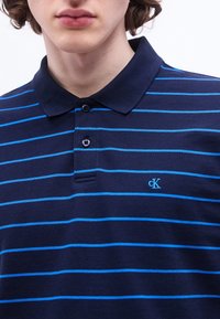 Jeune personne portant un polo bleu marine avec des rayures horizontales bleu clair et un petit logo bleu "cK" sur la poitrine gauche.
