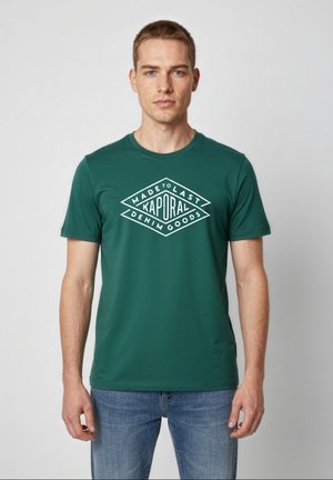 Mężczyzna w zielonym t-shircie Kaporal z nadrukiem „Made to Last Denim Goods”, w połączeniu z niebieskimi dżinsami, stoi na jednolitym tle.