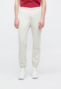 Pantalones deportivos beige claro con puños y cintura elásticos, hechos de material suave, combinados con zapatillas deportivas blancas y una camiseta roja.