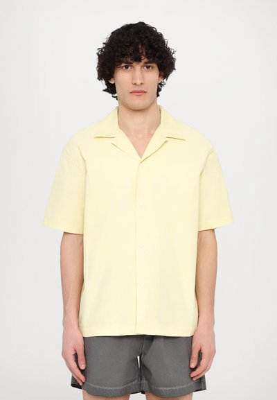 Camisa de manga corta, con botones, en tela amarilla pastel con cuello muesca y textura suave, combinada con pantalones cortos grises.