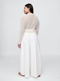 Kremfarget, åpen heklet cardigan med lange ermer og scalloped krage, sammen med vide, hvite bukser og flate sandaler.