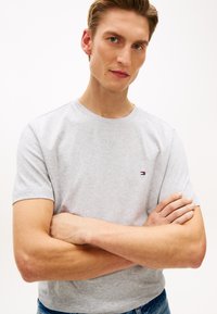 Γκρι κοντομάνικο μπλουζάκι από βαμβάκι με στρογγυλή λαιμόκοψη. Διαθέτει μικρό λογότυπο στο αριστερό στήθος. Το μοντέλο έχει σταυρωμένα τα χέρια.