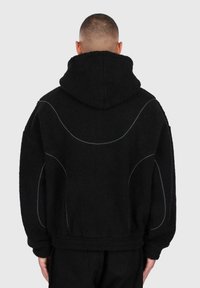 Uomo visto di spalle che indossa una giacca in pile con cappuccio nera, con dettagli di piping riflettente curvo sulla schiena e sulle maniche.