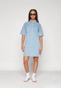 Lyseblå denim skjortekjole med korte ermer, knapfront og brystlomme. Har vertikale hvite striper og en avslappet passform. Brukt med hvite joggesko.