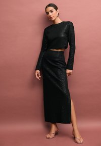 Black sequined two-piece outfit featuring a cropped top with long sleeves and a high-waisted skirt with a side slit. Transparent heeled sandals.