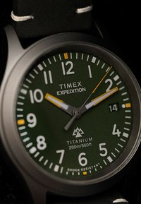 Timex Expedition sat sa titanskom kućištem, zelenim brojčanikom, bijelim i žutim oznakama sati te crnim remenom. Vodootporan do 200 m, otporan na udarce.